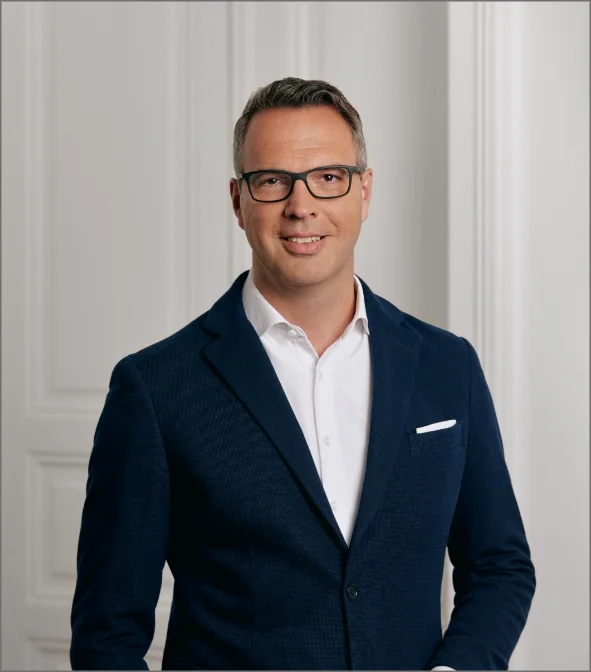 Porträt eines lächelnden Mannes mit Brille im dunkelblauen Sakko vor heller Wand